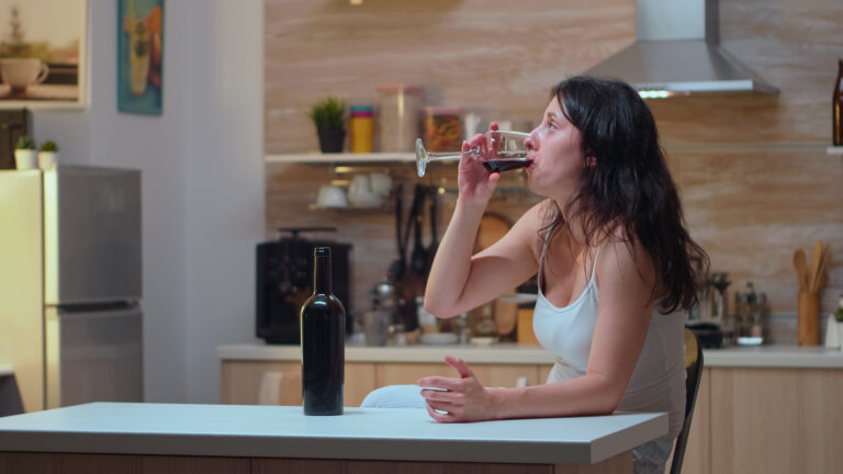 Woman drinking red wine in kitchen, illustrating personal wellness challenges.