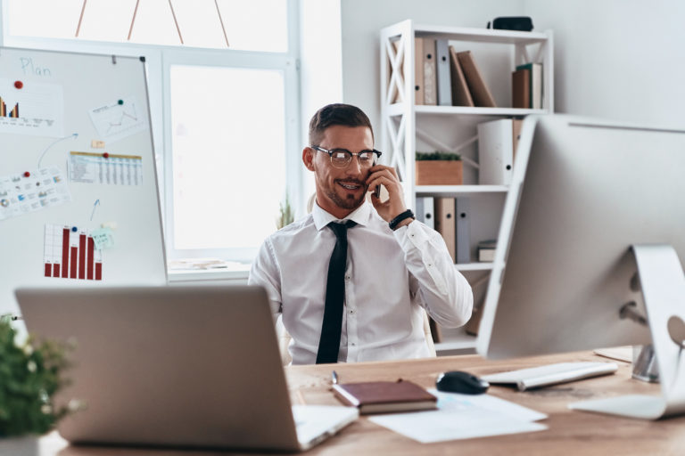 a person in a white shirt and tie sitting at a desk with a laptop and phone