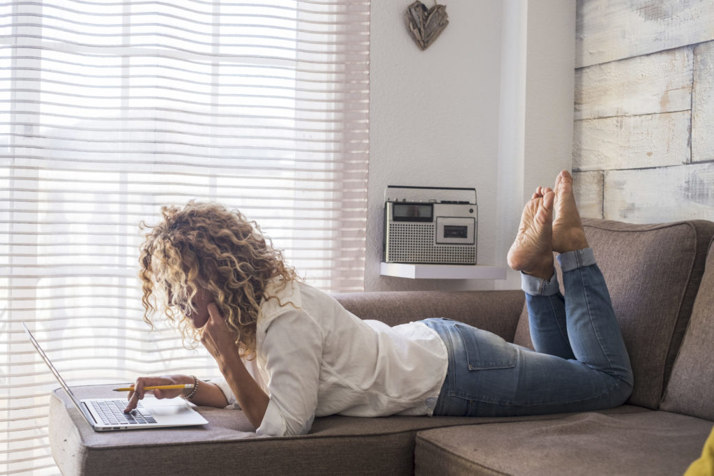 Curly woman at home lay down and use computer laptop on the sofa in reverse position